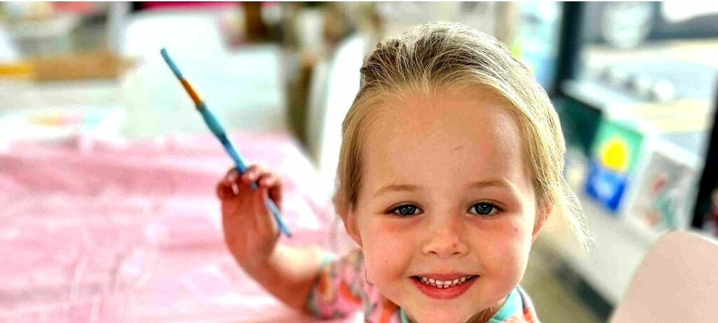 A young girl with freckles and blond hair looking straight into the camera and smiling, while holding a paintbrush